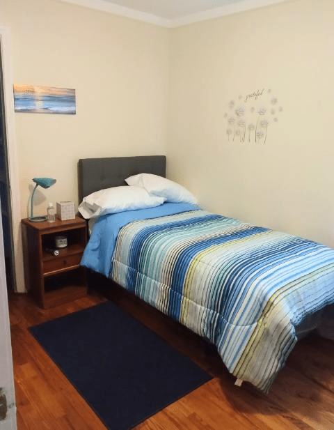 A cozy bedroom with a single bed featuring blue striped bedding, a nightstand with a lamp, and decorative wall art.