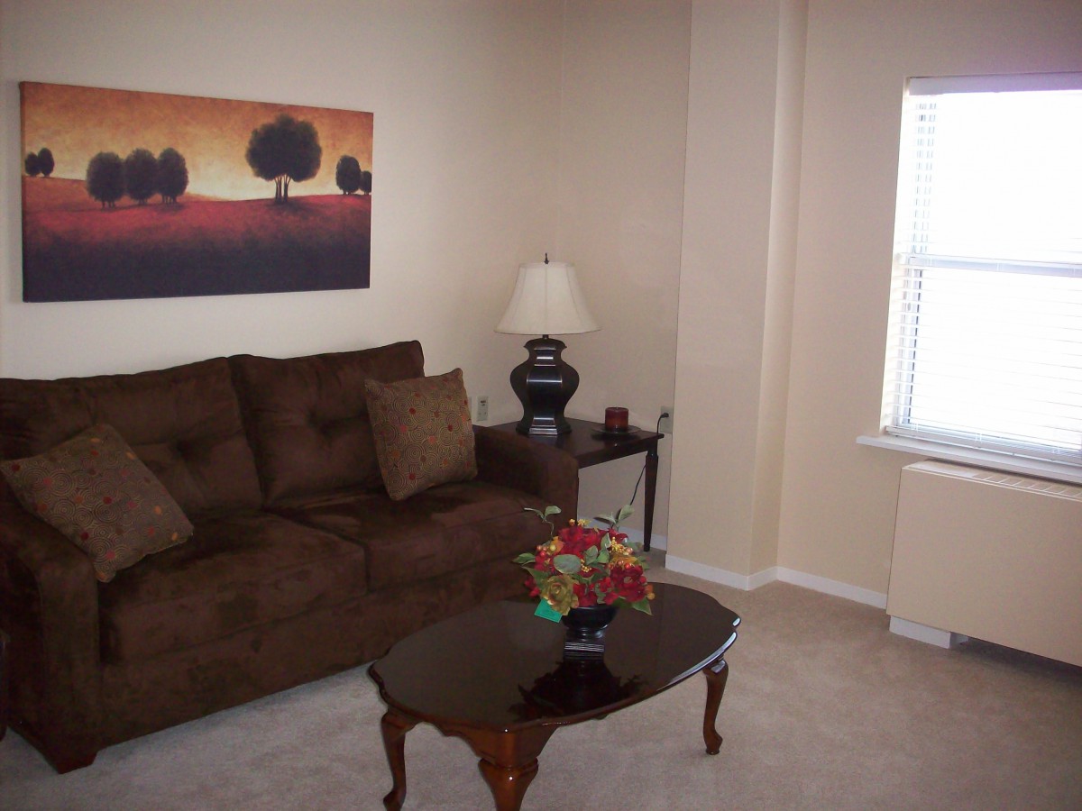 A cozy living room with a brown sofa, a lamp on a side table, and a coffee table in front of the sofa. A large painting hangs on the wall above the sofa.