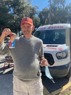 A man wearing a cap stands outdoors holding ice cream in front of an ice cream van on a sunny day.