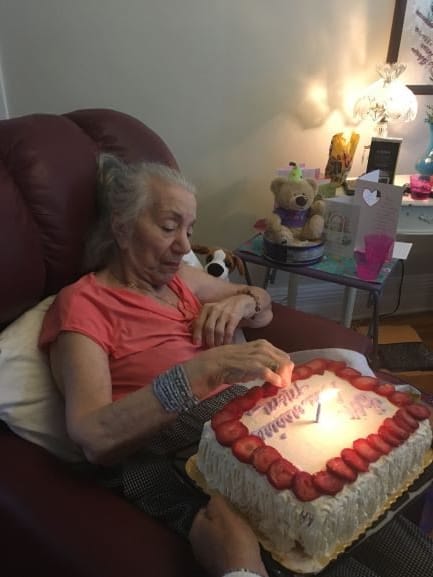 An elderly woman sitting in a chair prepares to blow out candles on her birthday cake.