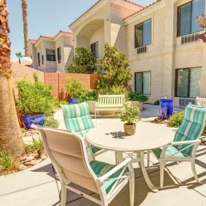A sunny patio area with a table and chairs, surrounded by garden plants, adjacent to an apartment building.