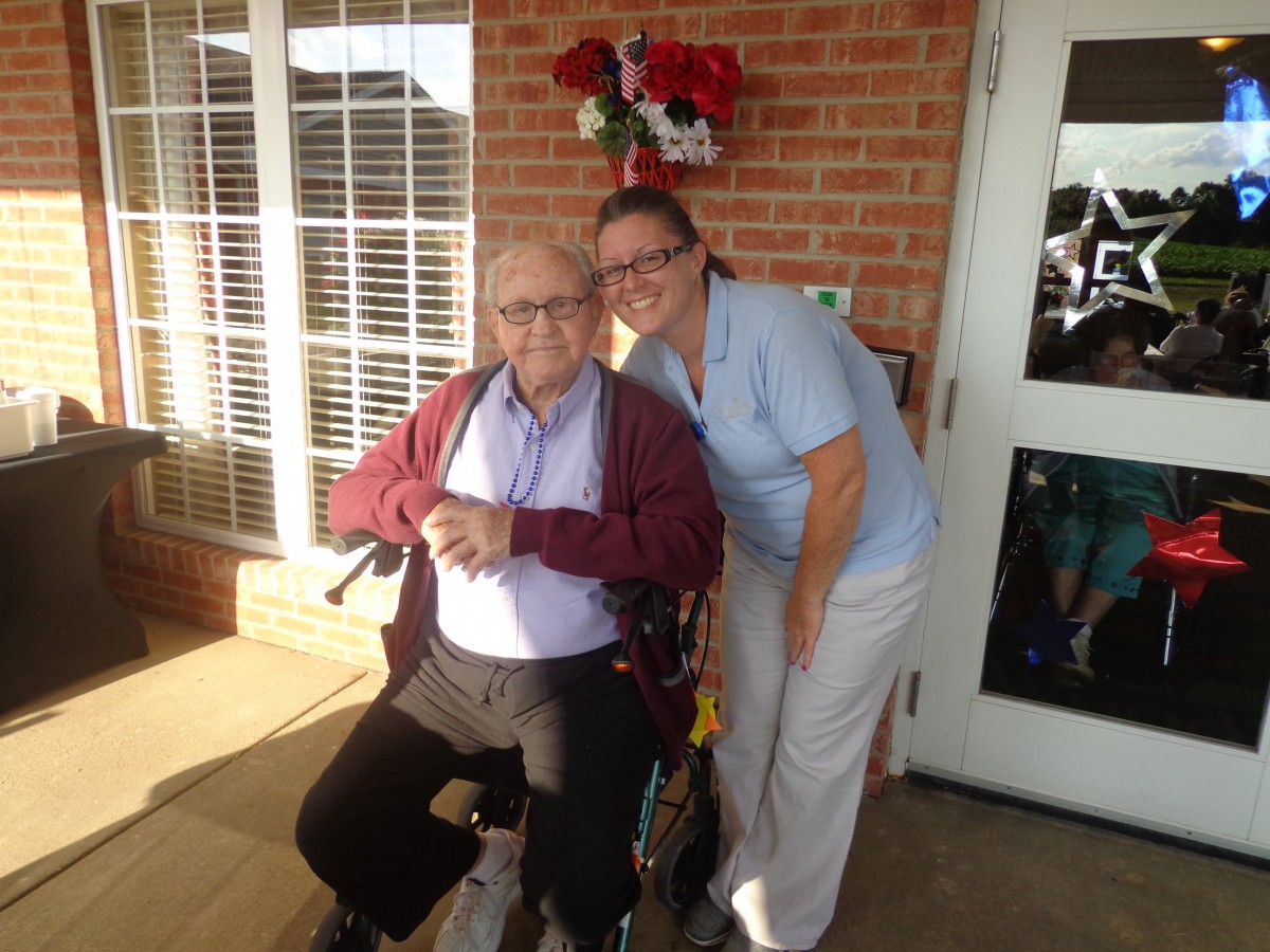 An elderly man in a wheelchair with a caregiver smiling beside him outside a building.