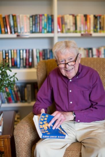 An elderly man wearing glasses sits in an armchair, smiling while holding a book in a library setting.