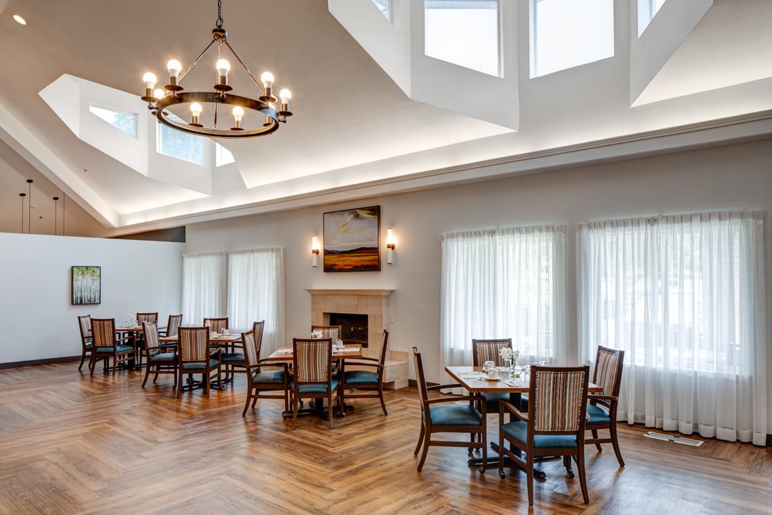 A bright and elegant dining room with multiple tables, a central chandelier, and large windows allowing plenty of natural light.