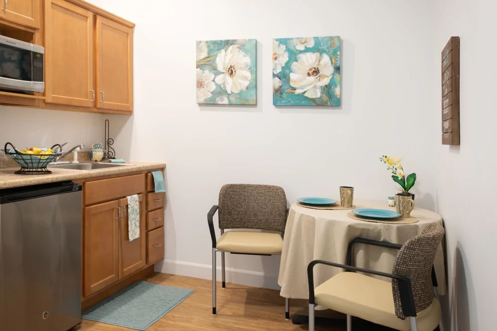 A cozy kitchen and dining area with floral artwork, a small table set for two, and warm wooden cabinetry.