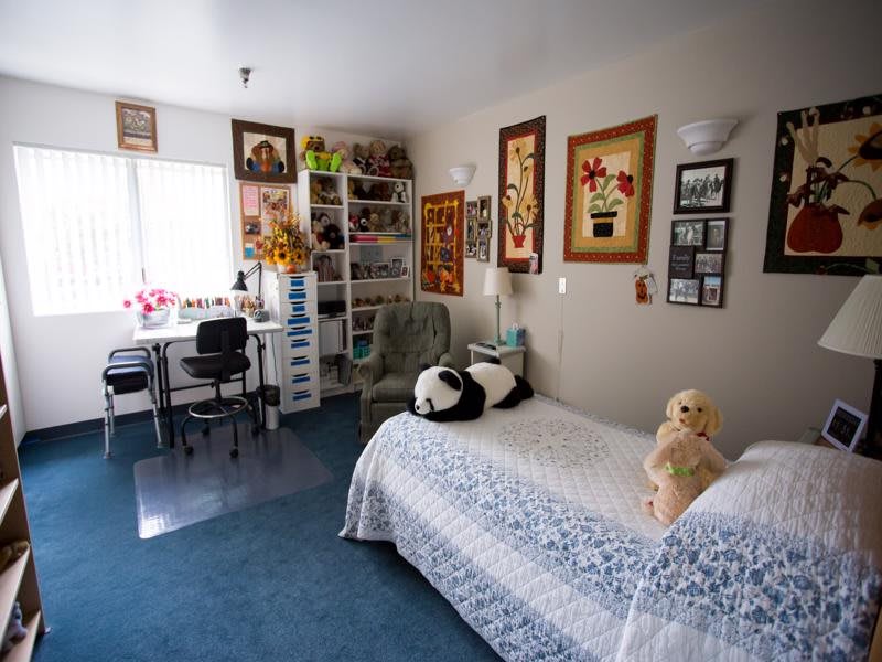 A cozy bedroom with artwork on the walls, a bed with plush toys, and a study area by the window.