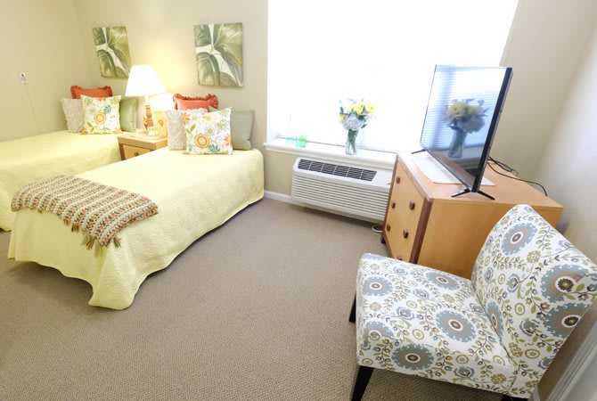 A cozy bedroom with a neatly made bed, a patterned chair, and a dresser with a television.
