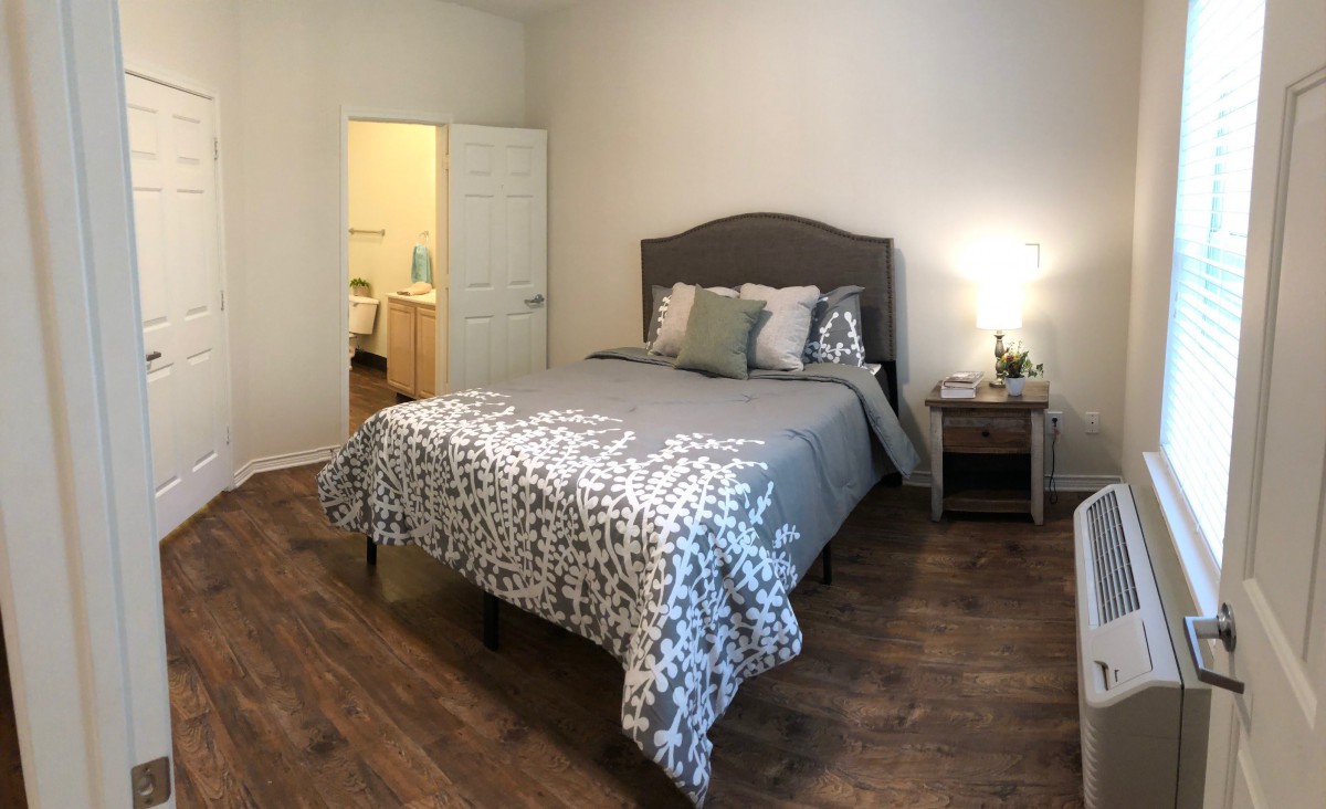 A cozy bedroom with a neatly made bed, nightstand, and lamp on a hardwood floor.