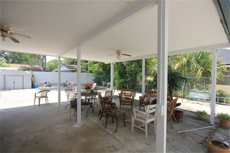 A spacious outdoor patio with various seating arrangements under a covered roof, featuring a ceiling fan and surrounded by greenery.