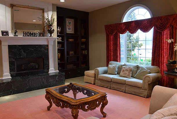 A cozy living room features a plush sofa, an ornate coffee table, and a fireplace adorned with decor. Natural light pours in through a large window framed by red curtains.