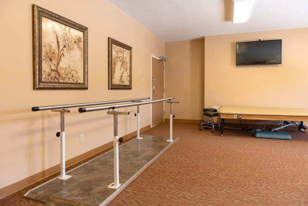 A well-lit rehabilitation room featuring parallel bars for physical therapy, decorated with wall art and equipped with a television.