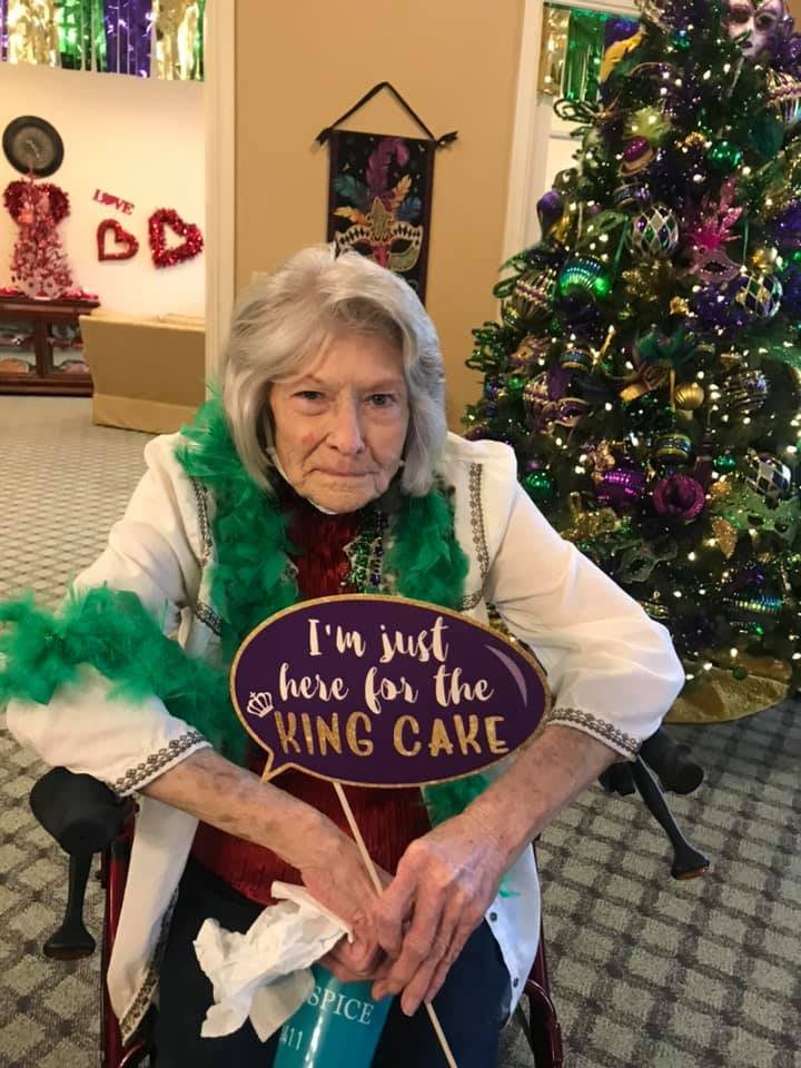 An elderly woman seated in front of a decorated Christmas tree, holding a sign that reads 'I'm just here for the King Cake'.
