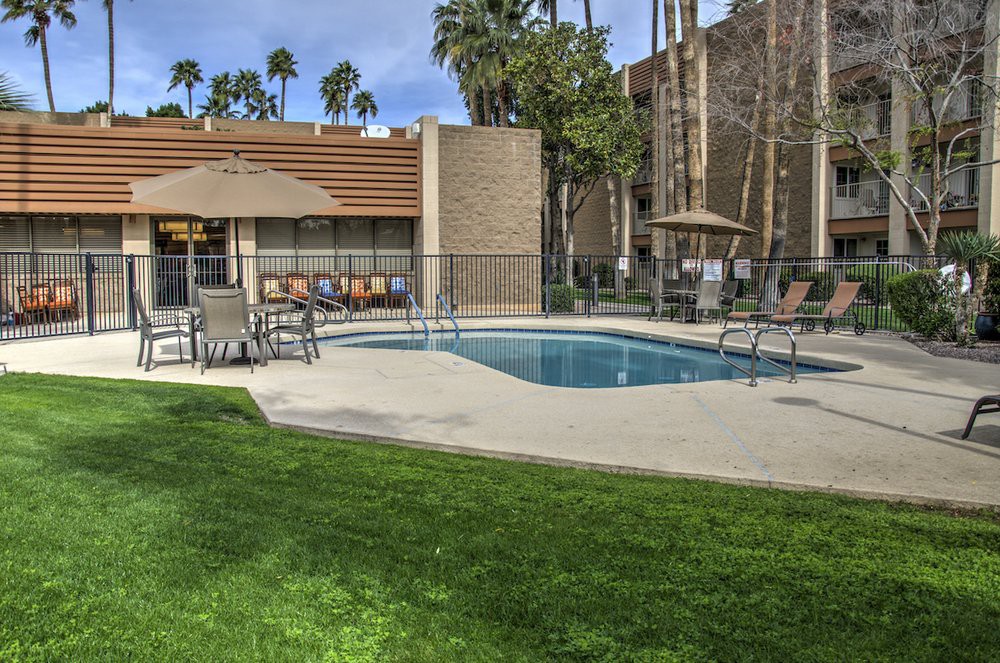 An outdoor swimming pool surrounded by patio furniture and palm trees near a residential building.