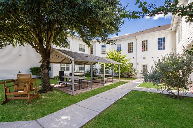 A peaceful garden area with a shaded patio and seating in front of a building.