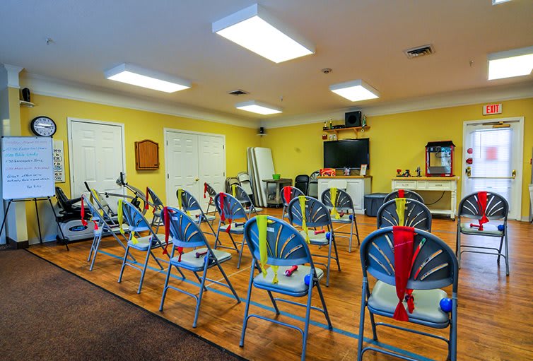 A well-lit classroom with rows of blue chairs, a television on the wall, and bright yellow walls.