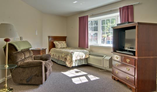 A cozy bedroom featuring a single bed, television, and large window with curtains.