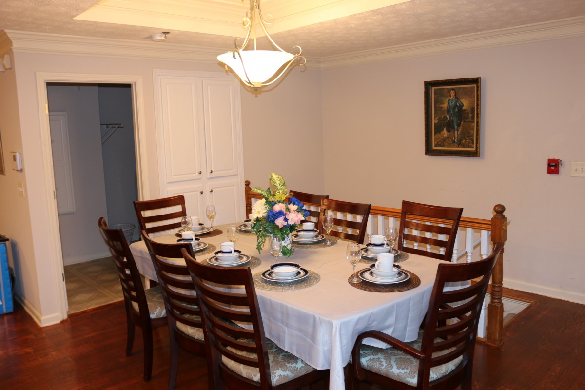 A neatly arranged dining room with a table set for eight, featuring a chandelier above and decorative flowers in the center.