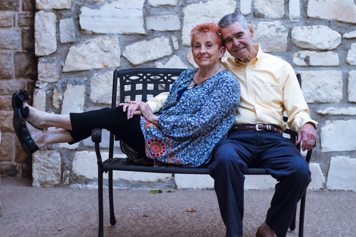 An elderly couple sitting together on a bench in front of a stone wall.