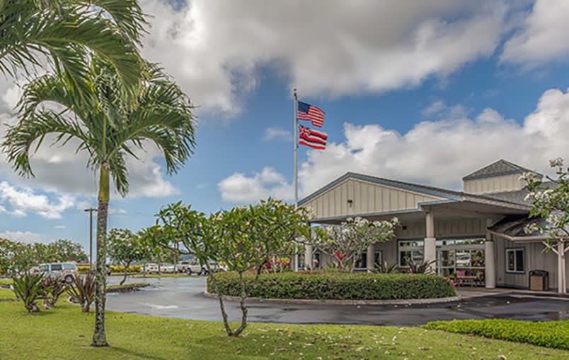 A building with an American flag flying outside, surrounded by palm trees and greenery under a cloudy sky.