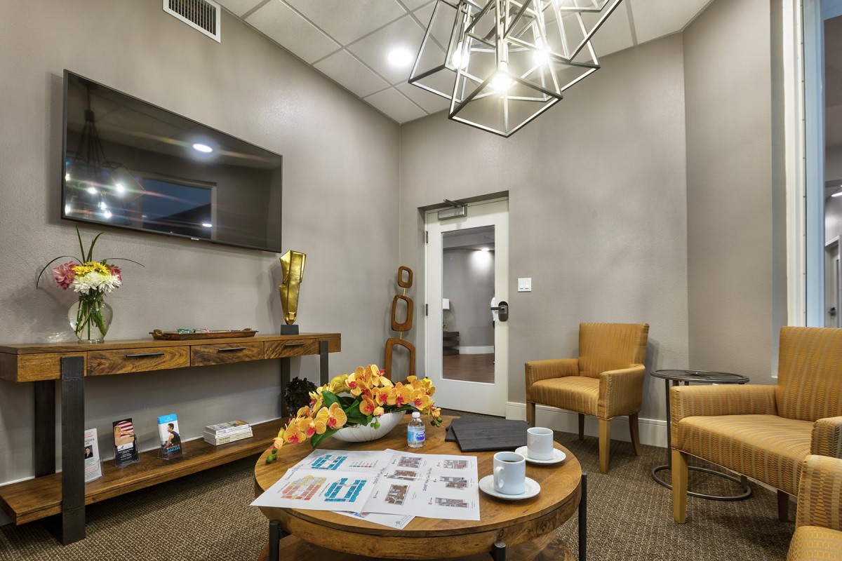 A modern waiting room featuring comfortable chairs, a coffee table with magazines and cups, a television, and stylish lighting.