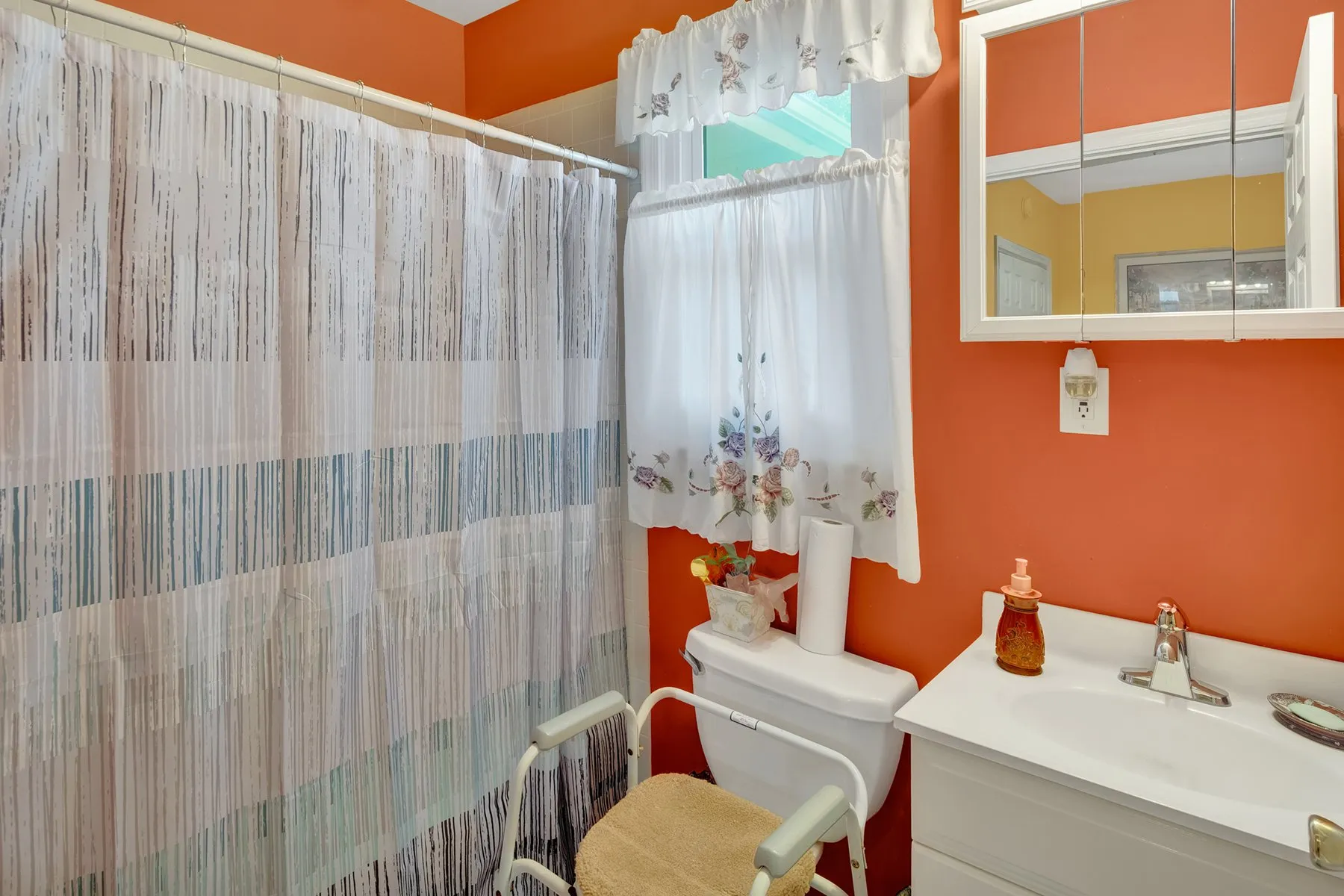 A cozy bathroom with vibrant orange walls, featuring a shower curtain and a small window with lace curtains.