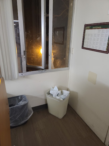 A corner of an office room featuring a window, a trash can filled with paper, and a wall calendar.