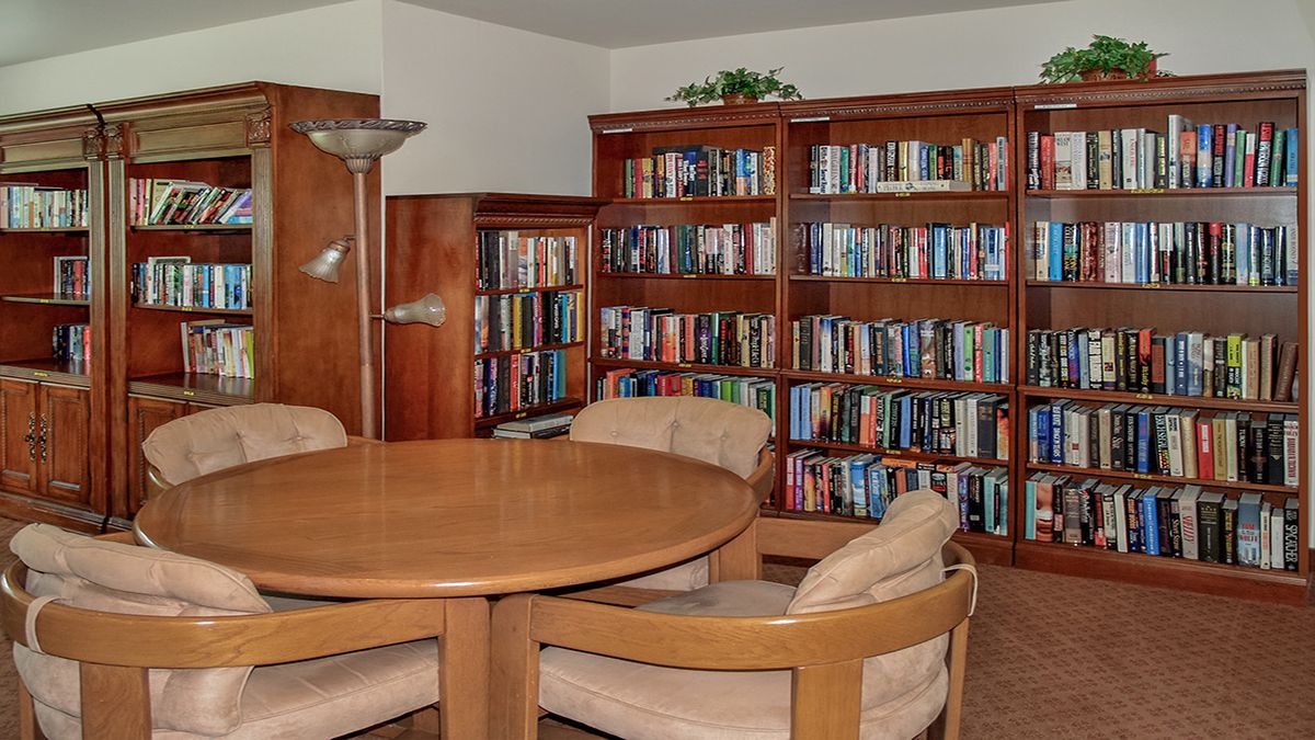 A cozy library room featuring wooden bookshelves filled with books, a round wooden table, and cushioned chairs.