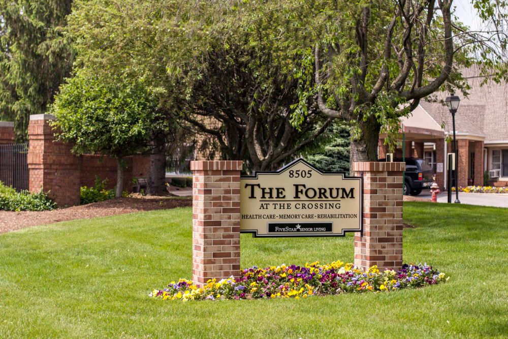 A sign for 'The Forum at the Crossing' surrounded by flowers and trees, marking the entrance to a building complex.