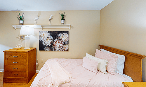 A cozy bedroom with a neatly made bed, decorative artwork, and a small plant on a shelf.