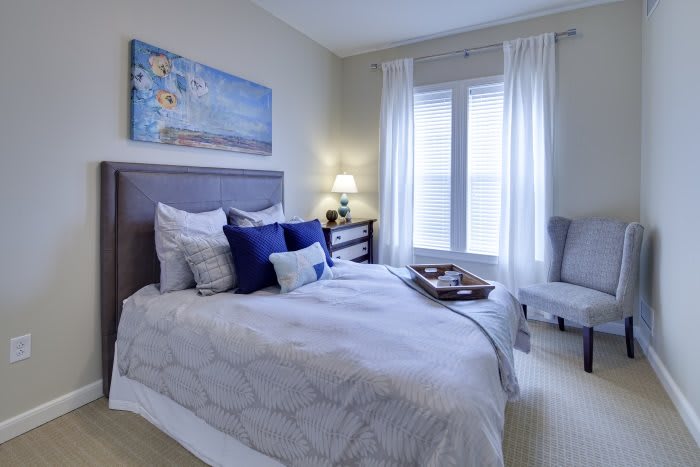 A cozy bedroom interior featuring a neatly made bed with decorative pillows, a bedside lamp, and a piece of artwork above the headboard. Natural light streams in through a large window with white curtains.
