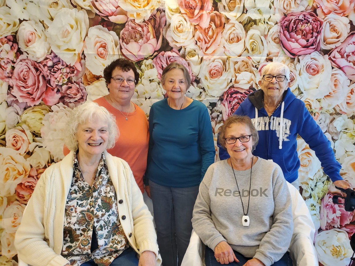 A group of five elderly women posing together in front of a floral backdrop.