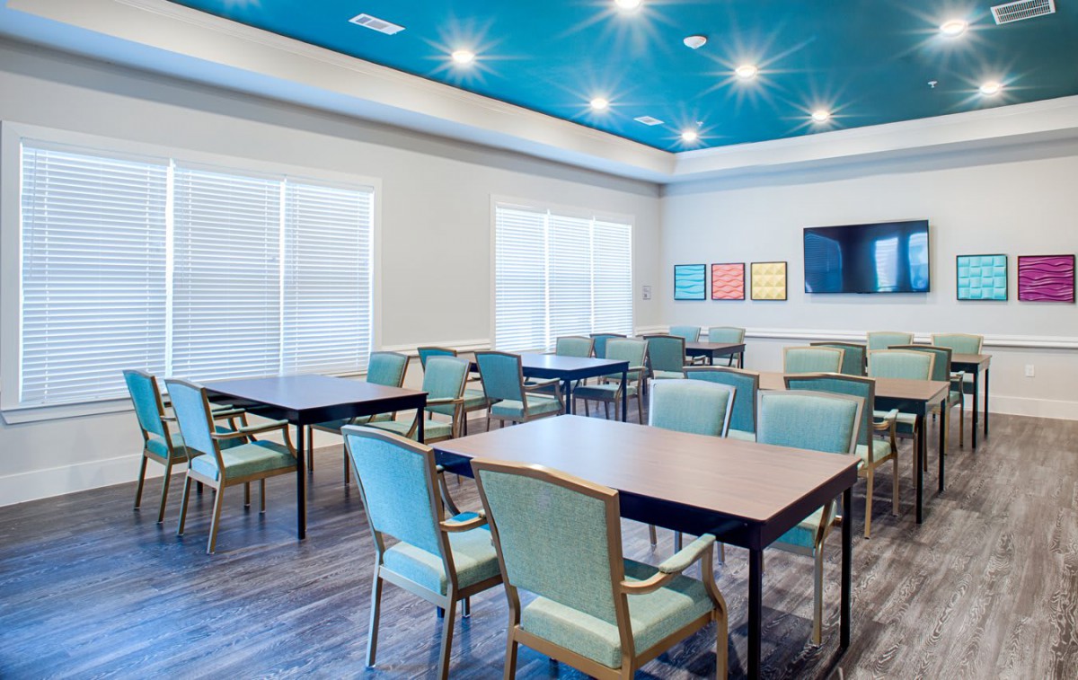 A modern meeting room with multiple tables and chairs, featuring bright lighting and a television on the wall.