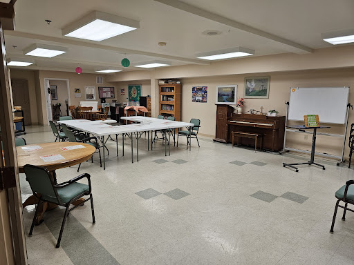 A spacious community room set up with tables and chairs, featuring a piano and artwork on the walls.