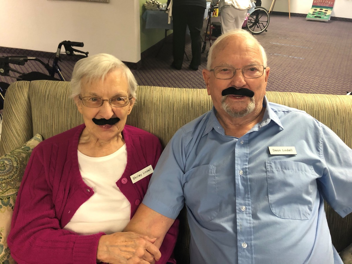An elderly couple sitting together with playful fake moustaches.