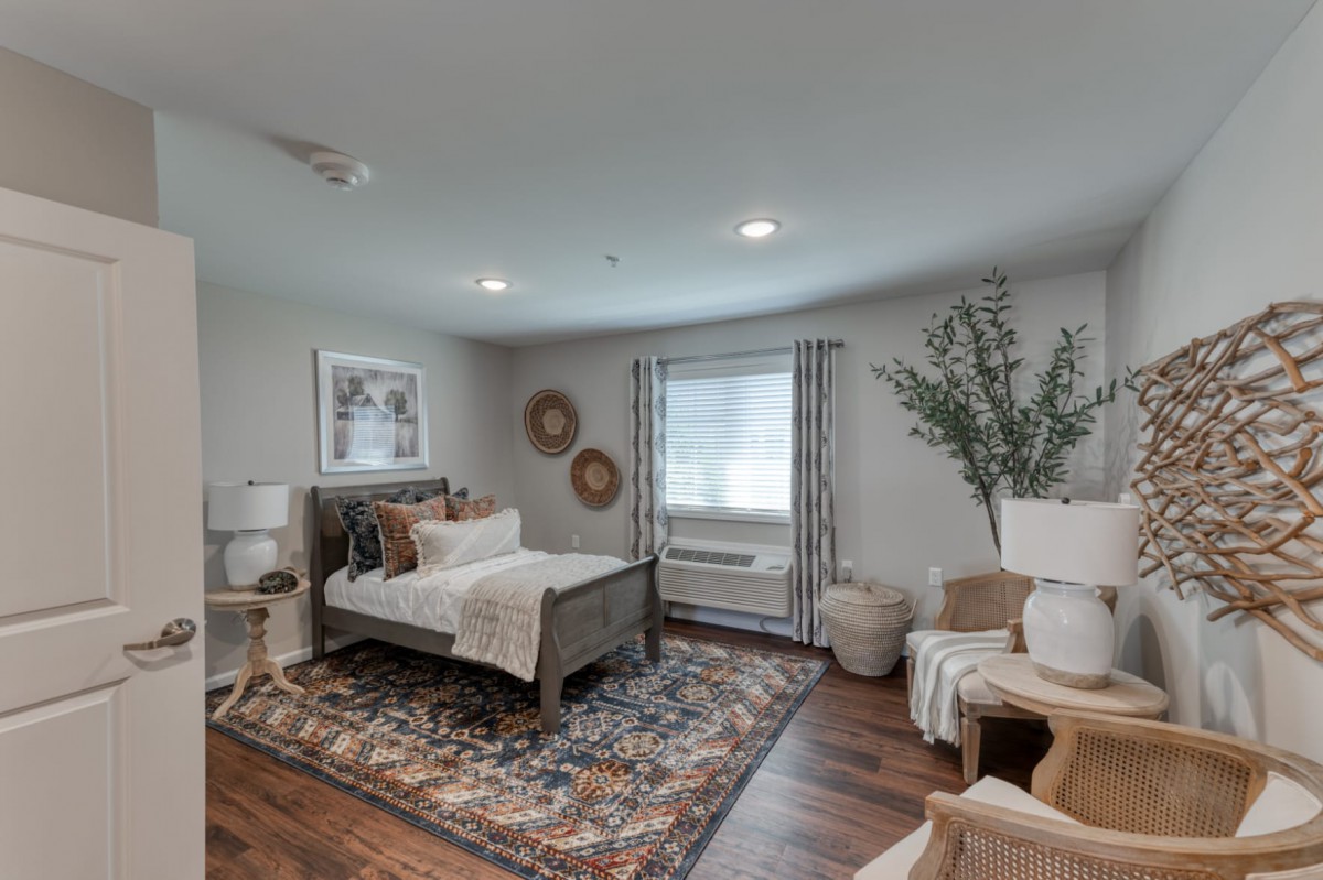 A modern bedroom with neutral tones featuring a patterned rug, wooden flooring, and decorative elements like wall art and plants.