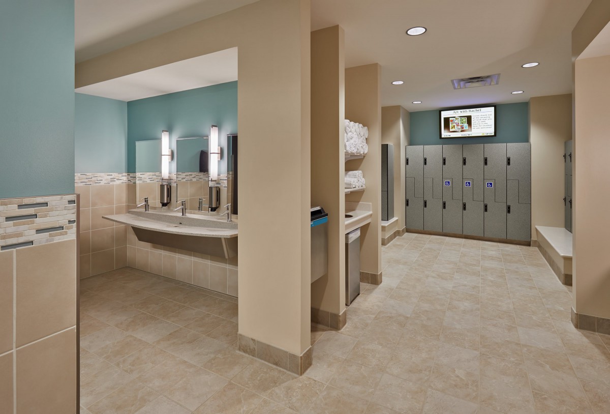 A modern locker room featuring a bathroom area with sinks and mirrors, alongside lockers and neatly stacked towels.