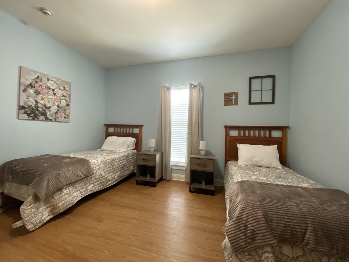 A cozy bedroom with two twin beds and a window between them, featuring light blue walls and wooden flooring.