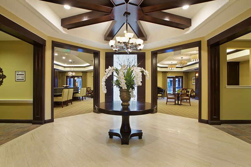A luxurious lobby featuring a central table with a floral arrangement under an elegant chandelier and wooden ceiling beams.