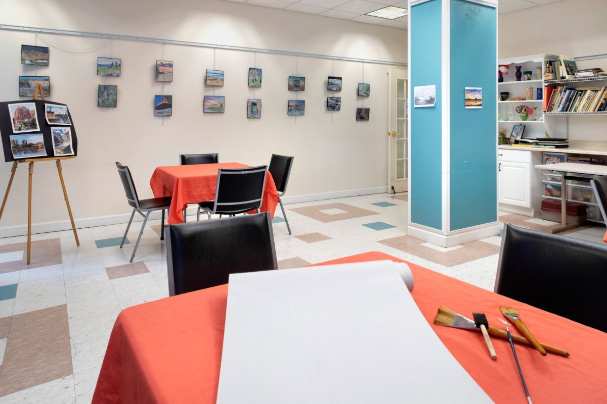 An art gallery interior with tables and chairs, featuring artwork displayed on the walls.