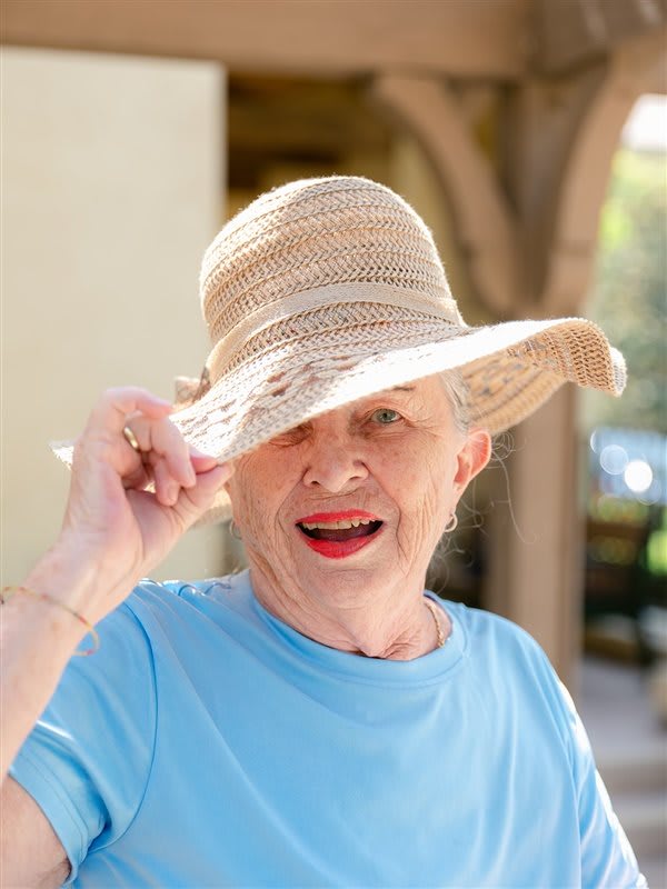 A smiling woman in a blue shirt holds her straw hat outdoors.