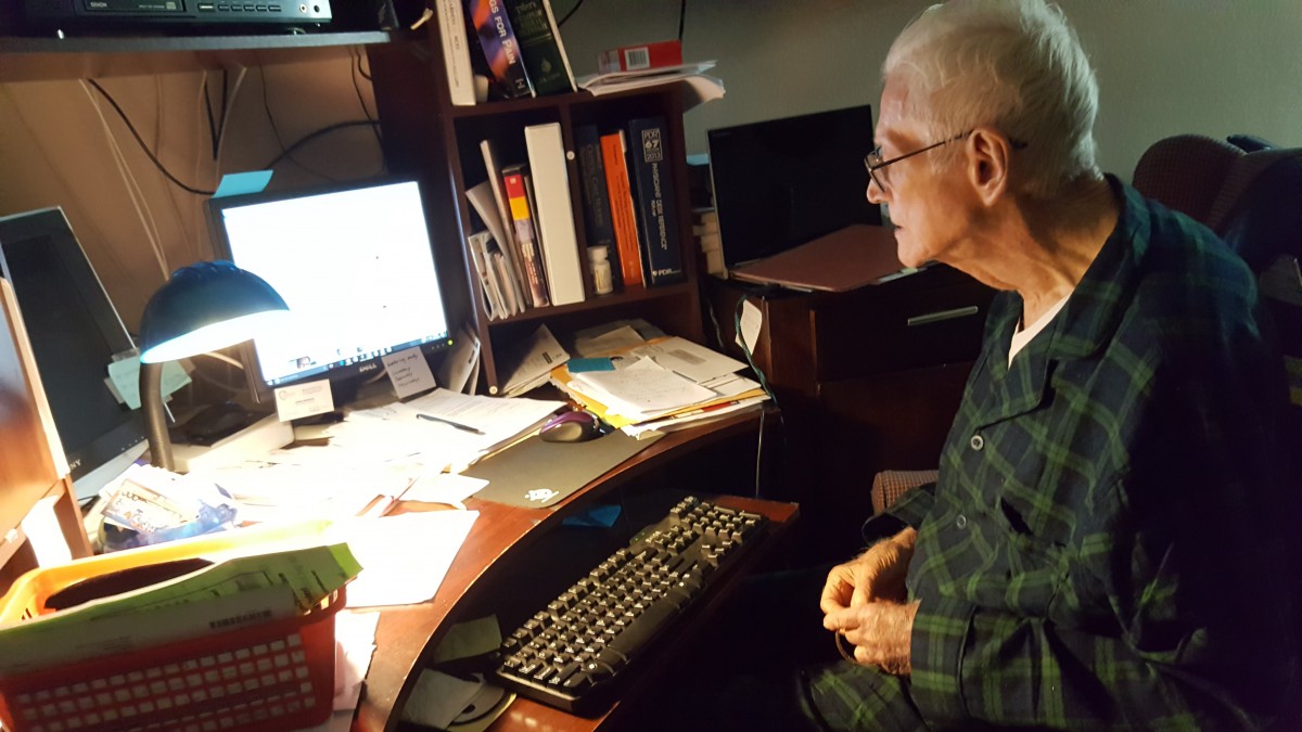 An elderly man sitting at a cluttered desk, working on a computer.