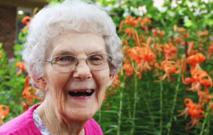 An elderly woman smiling in a garden with orange flowers in the background.