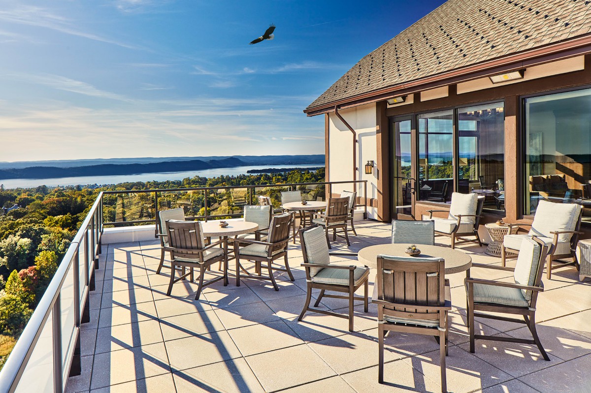 A scenic rooftop patio with tables and chairs overlooking a beautiful lake under a clear blue sky.