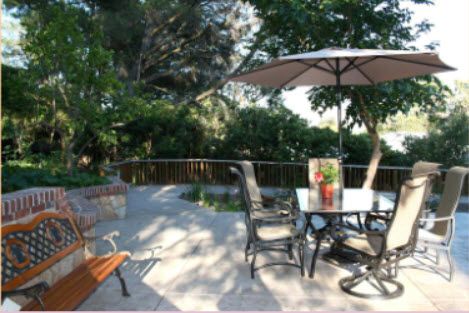 A sunny patio with a table, chairs, and an umbrella surrounded by lush garden greenery.