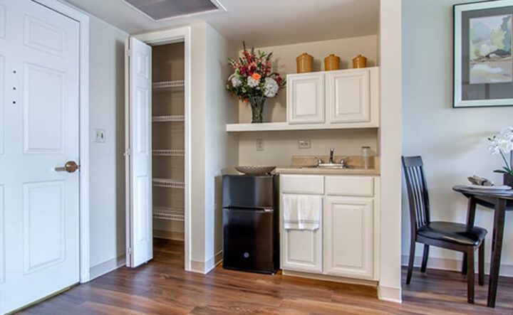 A small kitchenette with white cabinets, a mini fridge, and decorative flowers on the shelf.