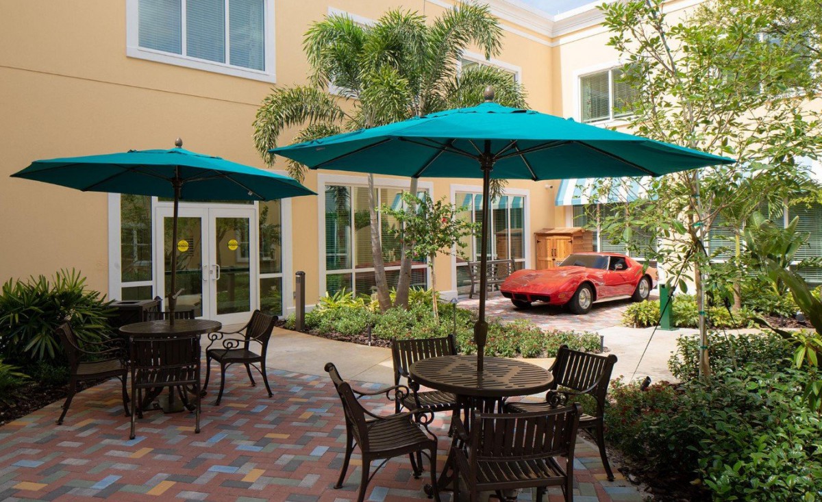 A pleasant outdoor patio area featuring tables, chairs, and umbrellas, with tropical plants and a red car parked nearby.