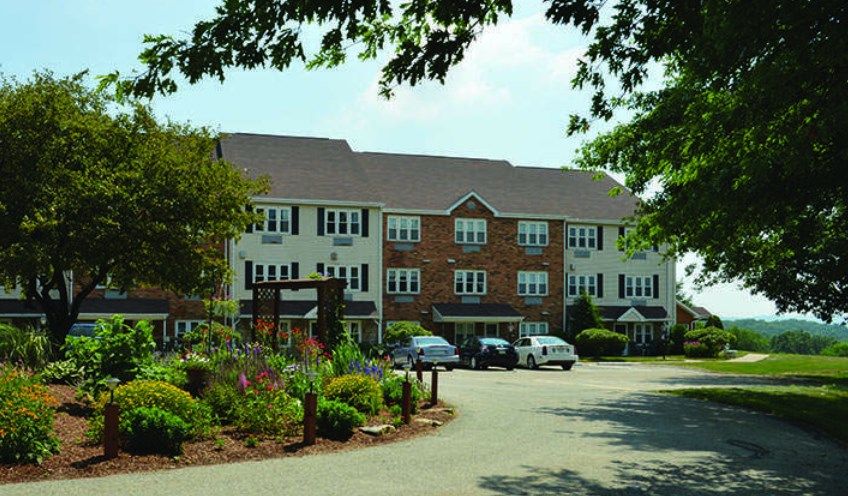 A three-story building with a parking lot surrounded by trees and landscaped gardens.