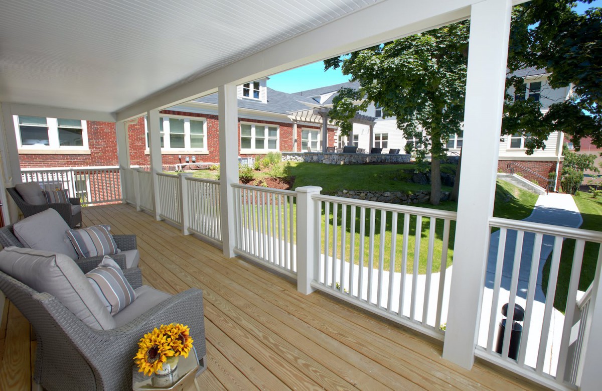 A cozy porch with a view of a garden and neighboring houses, featuring comfortable seating and decorative flowers.
