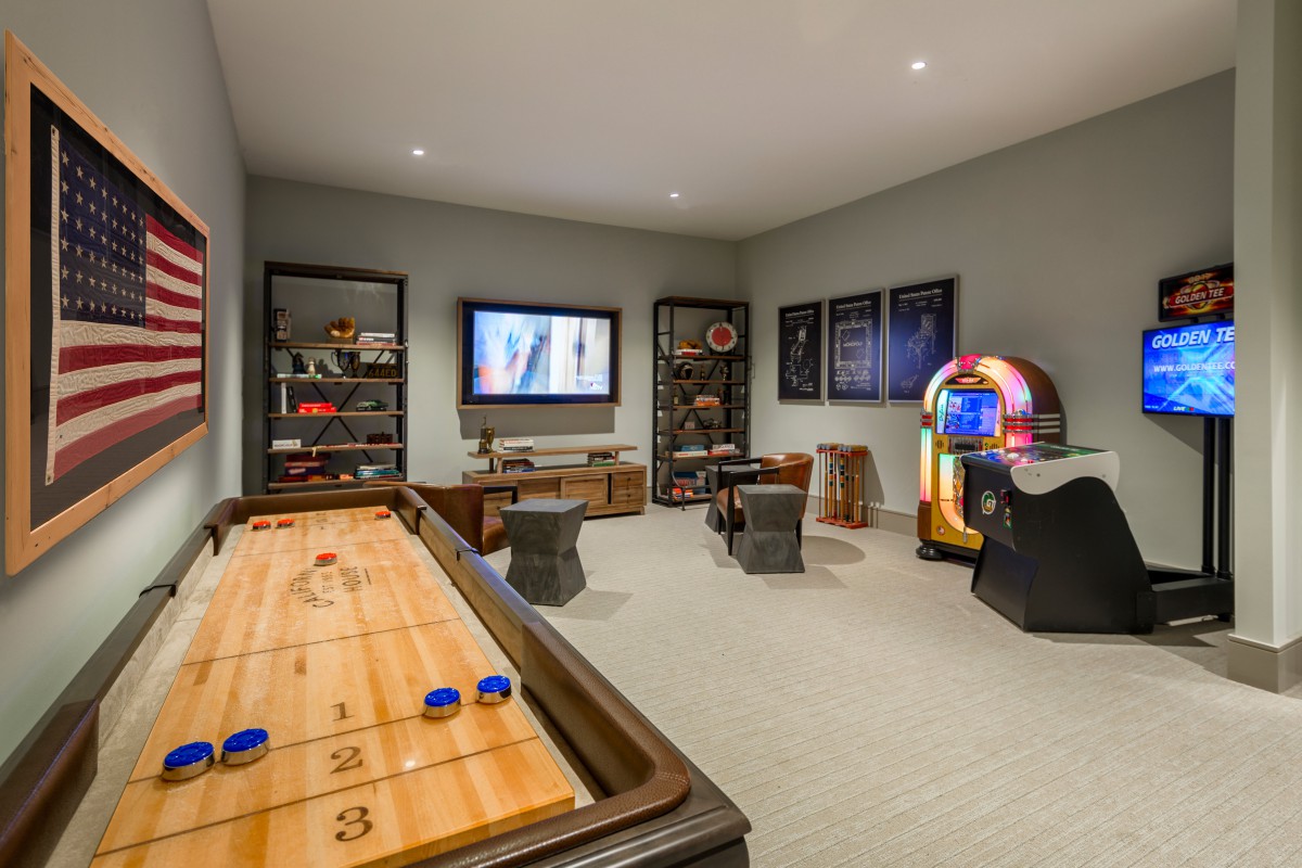 A comfortable game room featuring a shuffleboard table, vintage arcade machine, jukebox, and television with a cozy seating area.
