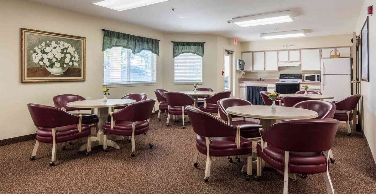 A cozy dining area with several round tables and cushioned chairs arranged neatly, under soft lighting and near large windows.